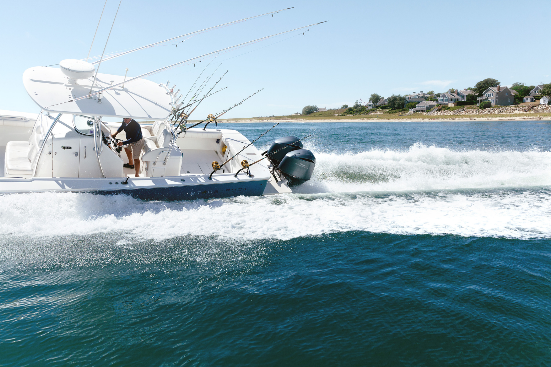 A boat speeding on water with fishing rods set, creating waves, and a shoreline with houses in the background.