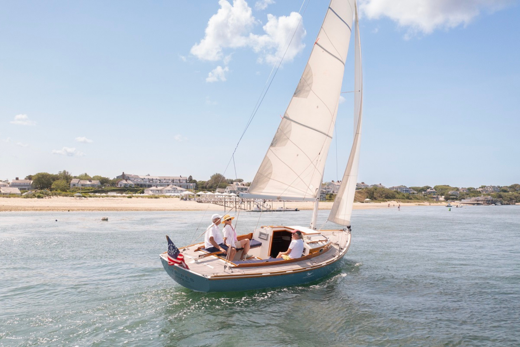 A sailboat with three people is cruising near a beach with buildings in the background under a partly cloudy sky.
