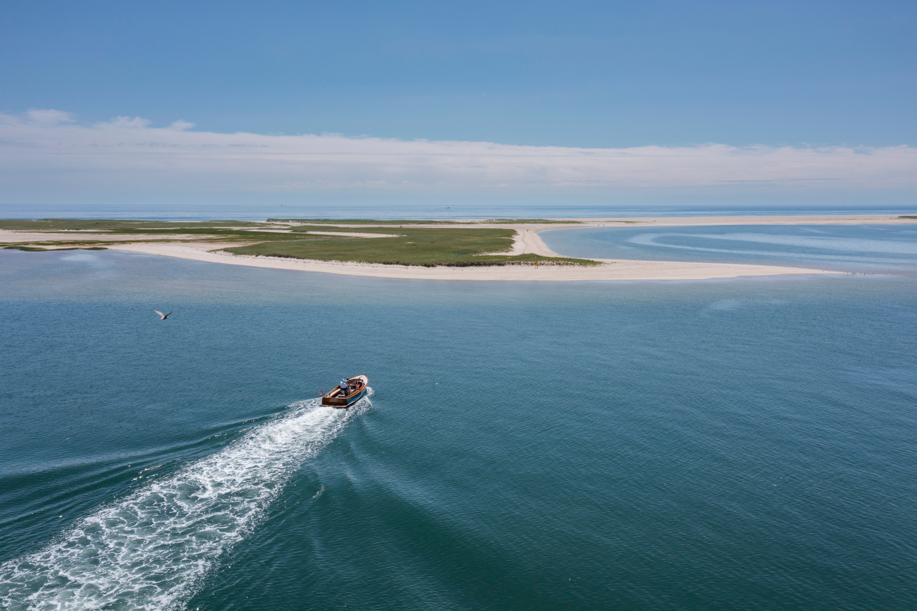 A small boat cruises on the blue ocean near a sandy island; a bird flies in the foreground under a clear sky.