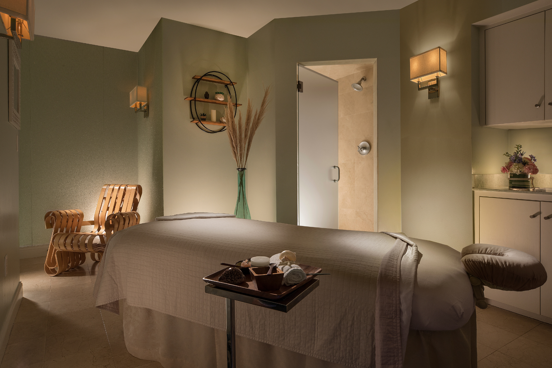 A serene spa room with a massage table, a wooden chair, soft lighting, and a glass vase with pampas grass, creating a calming ambiance.