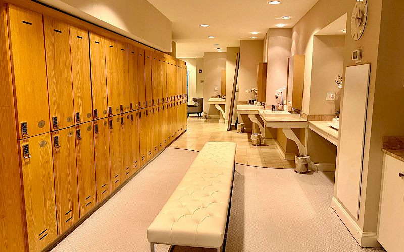 The image shows a locker room with wooden lockers, vanity stations, and a cushioned bench in a well-lit space.