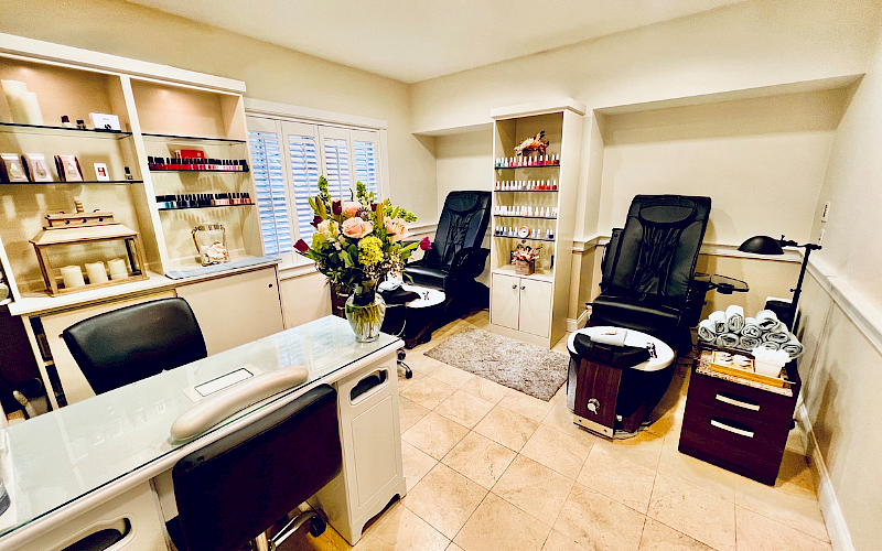 A cozy nail salon features manicure and pedicure stations, massage chairs, nail polish displays, and fresh flowers.