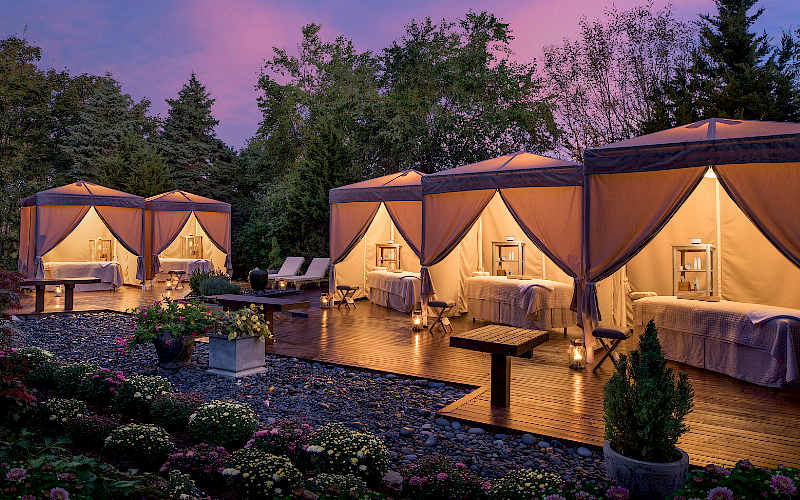 Outdoor cabanas with draped fabric, massage tables, and ambient lighting set in a garden at dusk.