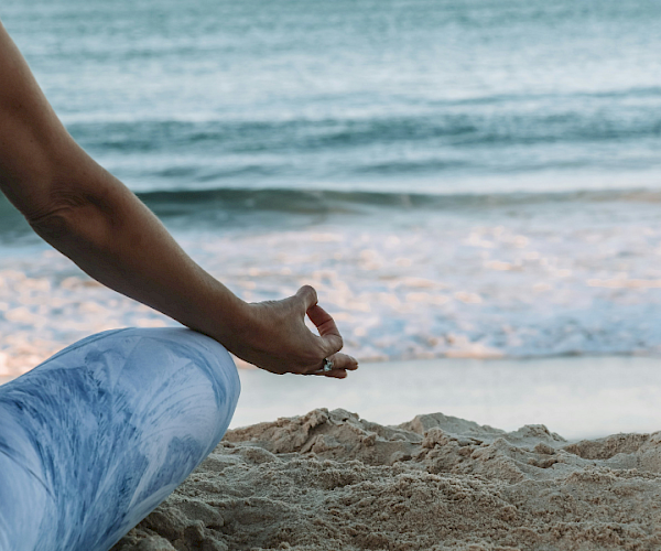 A person sits cross-legged on a sandy beach, meditating with a view of the ocean.
