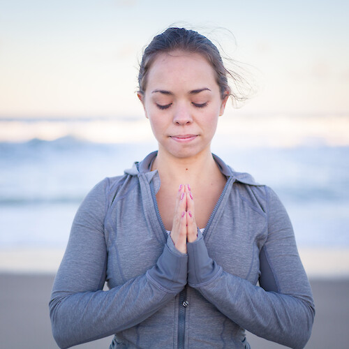 A person with closed eyes is practicing yoga or meditation on the beach, with hands in a prayer position, wearing a gray jacket.