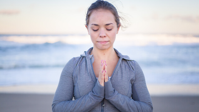 A person with closed eyes is practicing yoga or meditation on the beach, with hands in a prayer position, wearing a gray jacket.