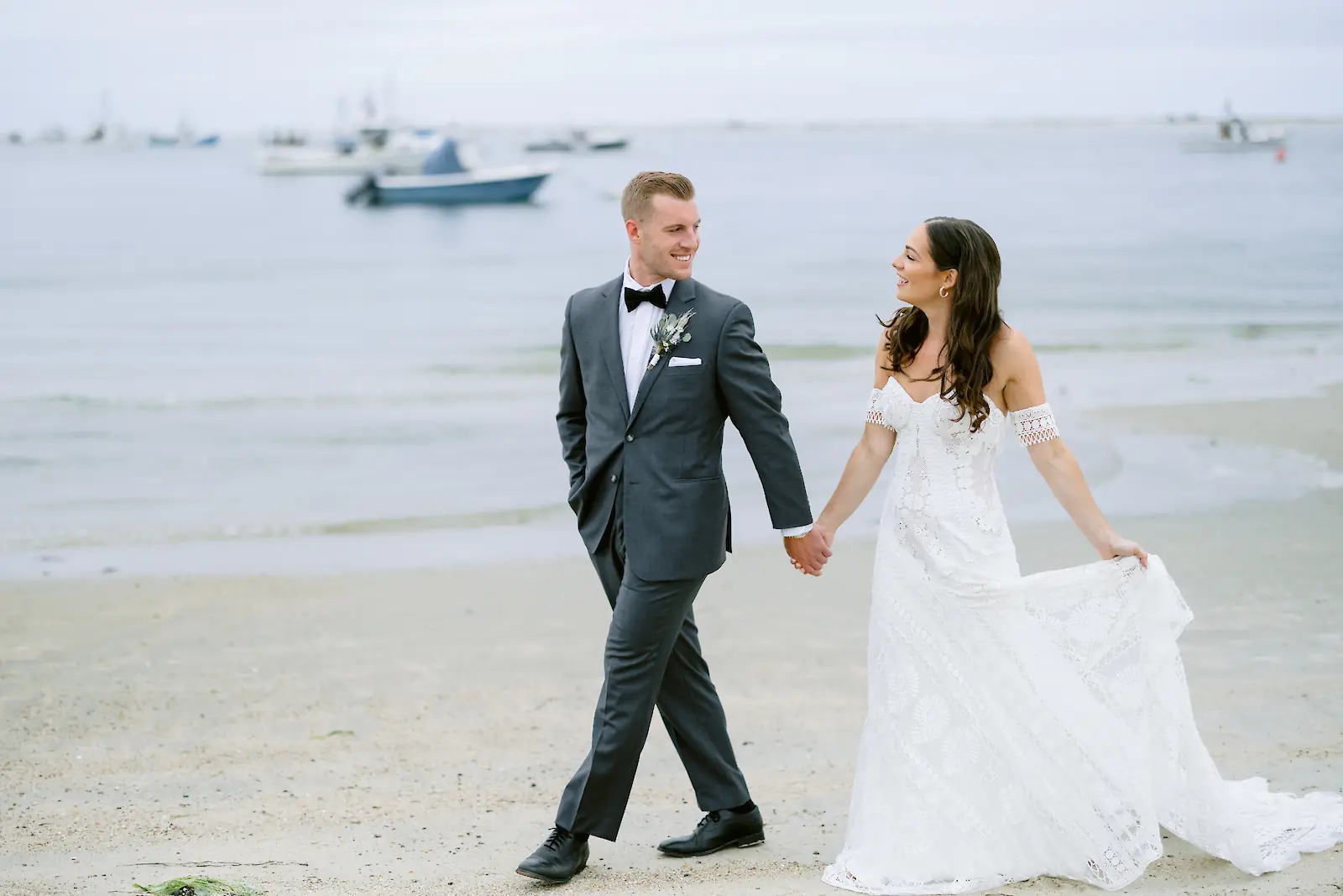A couple in wedding attire is walking hand in hand on a beach, with boats visible in the background.