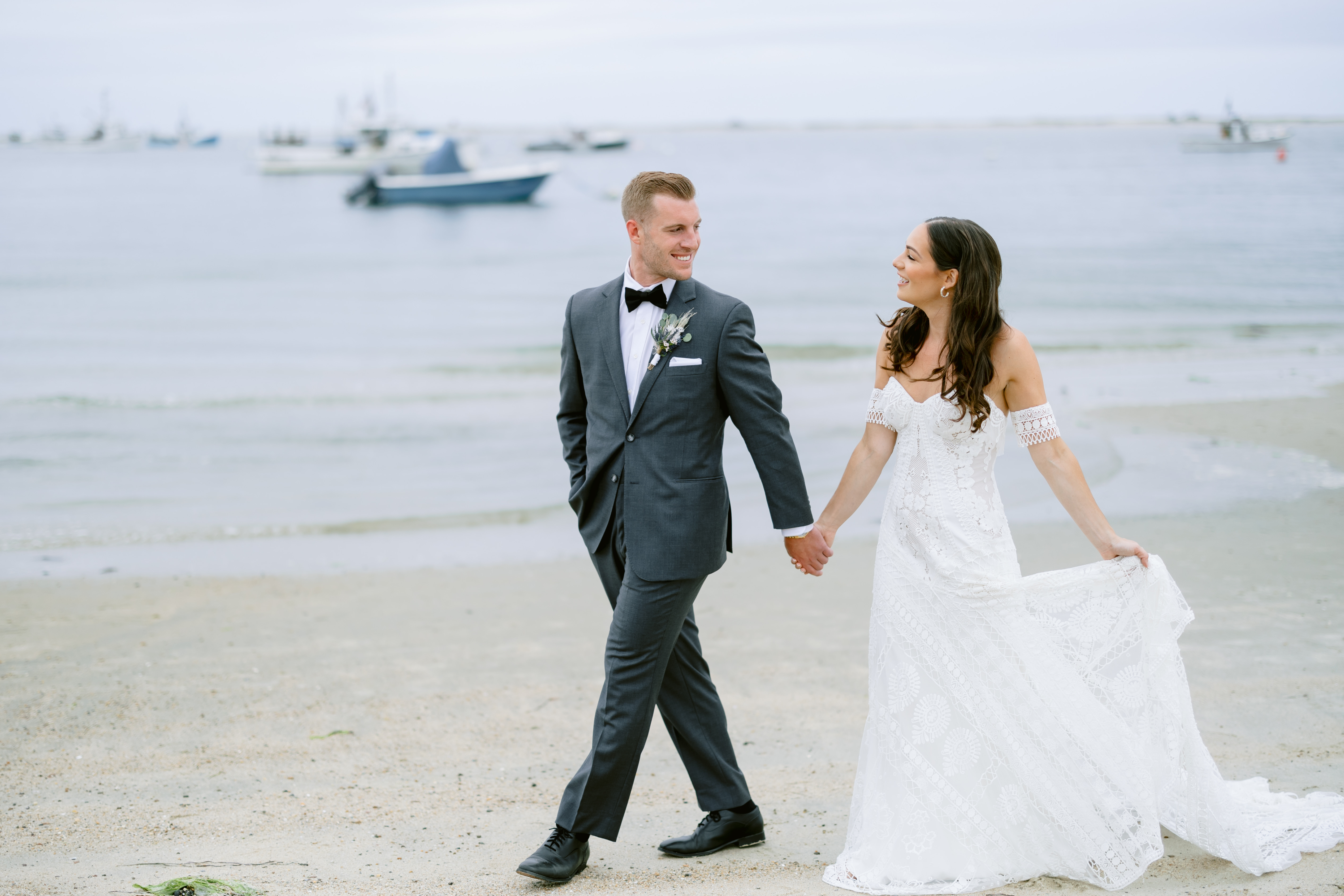 A couple in wedding attire is walking hand in hand on a beach, with boats visible in the background.