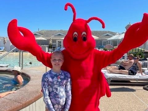 A person in a red lobster costume poses with a child near a pool, with people relaxing on lounge chairs in the background.