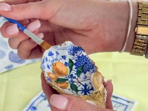 A person is painting a decorative seashell with intricate blue, yellow, and green patterns using a small brush.