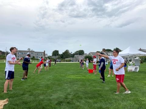 People are outside on a grassy field playing a game with a ball, wearing casual attire, with houses and trees in the background.