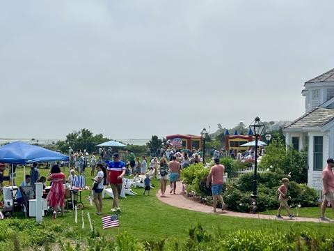 People enjoying a gathering outdoors with booths, inflatables, and American flags in a garden setting near a house.
