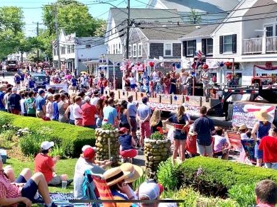 A festive outdoor event with a crowd, parade floats, and decorations, taking place in a quaint town setting.