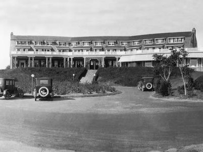 A vintage hotel building with a long facade, surrounded by parked antique cars and landscaped gardens, under a clear sky.
