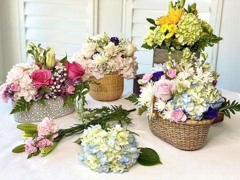 Assorted floral arrangements in baskets and vases on a table featuring roses, hydrangeas, daisies, and greenery.