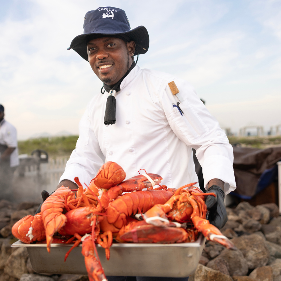 A chef in a white uniform and hat holds a tray of lobsters outdoors, with a scenic background.