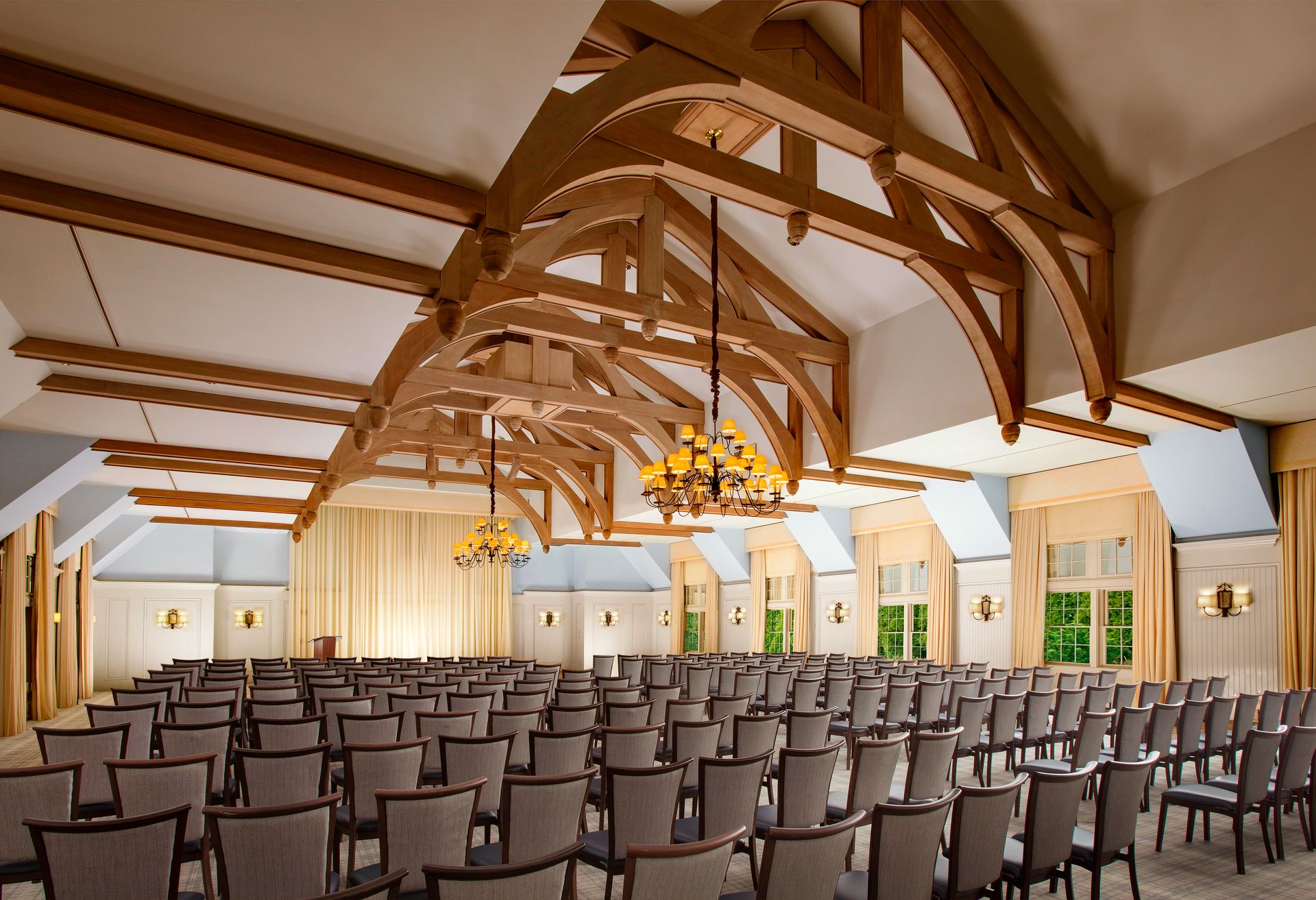 A spacious room with wooden ceiling beams, chandeliers, rows of chairs, and large windows, suitable for events or conferences.