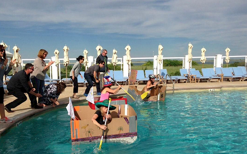 People are racing homemade cardboard boats in a pool, cheered on by onlookers by the edge of the water.