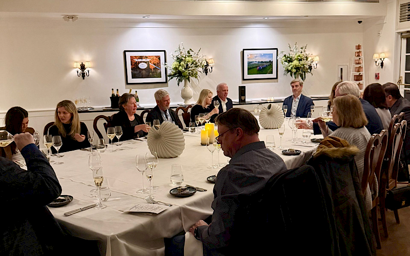 A group of people sit around a large, elegant dinner table with wine glasses and decorative centerpieces.