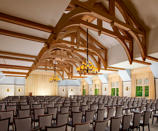 A spacious room with rows of chairs, wooden beams, chandeliers, and large windows, set for an event or presentation.