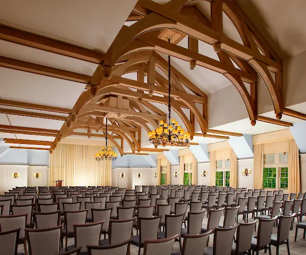 A spacious room with rows of chairs, wooden beams, chandeliers, and large windows, set for an event or presentation.