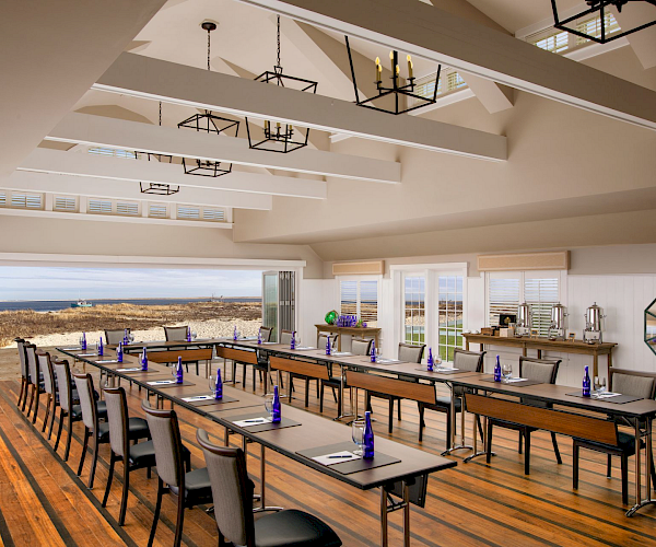 A bright conference room with tables and chairs set up. Blue bottles and notepads are on the tables, and there's an ocean view outside.