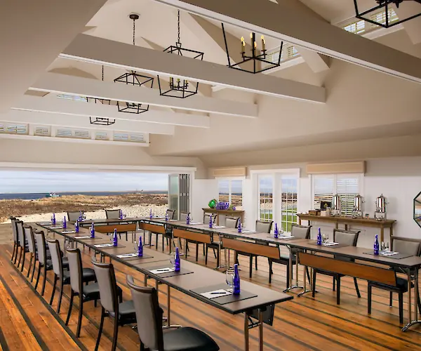 A bright conference room with tables and chairs set up. Blue bottles and notepads are on the tables, and there's an ocean view outside.