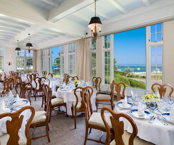 An elegant dining room with round tables set for a formal event, large windows showcasing a coastal view under a bright blue sky.