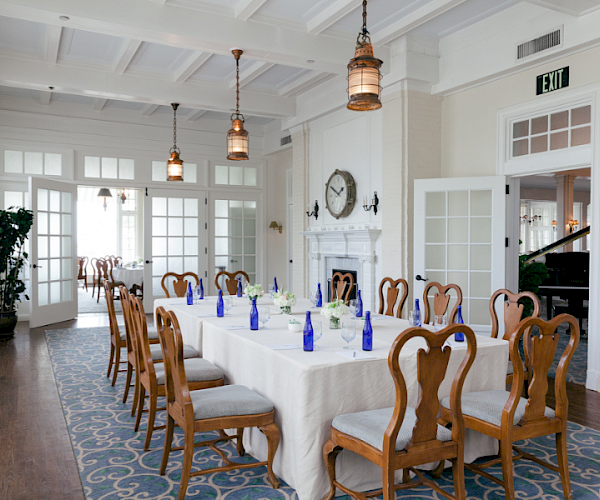 A well-set dining room with a large square table, wooden chairs, blue bottles, and elegant decor, featuring a wall clock and pendant lights.
