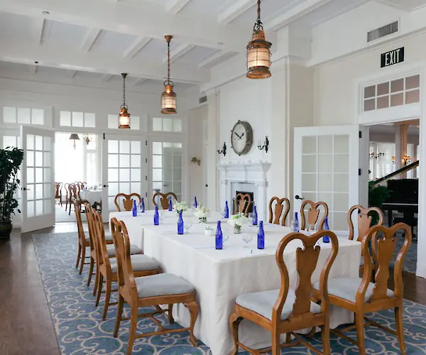 A well-set dining room with a large square table, wooden chairs, blue bottles, and elegant decor, featuring a wall clock and pendant lights.