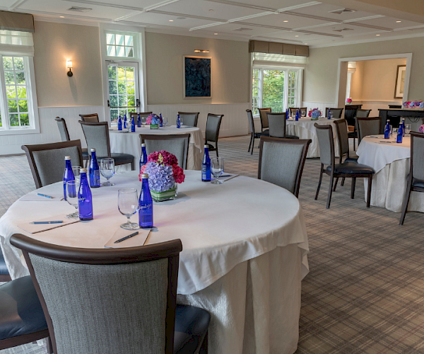 A room with round tables set for an event, featuring water bottles, glasses, and flower centerpieces on white tablecloths.
