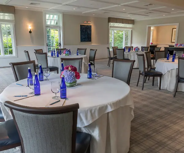 A room with round tables set for an event, featuring water bottles, glasses, and flower centerpieces on white tablecloths.