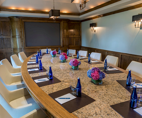 A conference room with a long marble table, arranged with notepads, pens, blue bottle water, glasses, and flowers, surrounded by chairs.