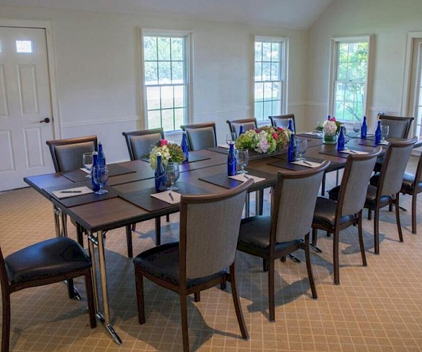 A meeting room with a long table, twelve chairs, blue water bottles, notebooks, pens, and floral centerpieces, set in natural light.
