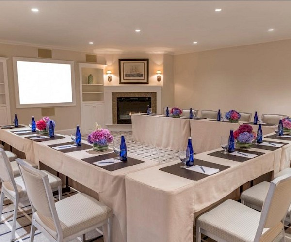 This image shows a U-shaped conference room setup with tables, chairs, notepads, blue bottles, and floral centerpieces, facing a screen.