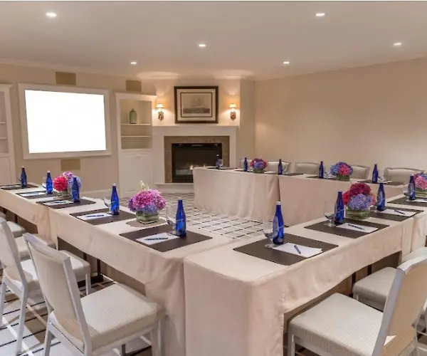 This image shows a U-shaped conference room setup with tables, chairs, notepads, blue bottles, and floral centerpieces, facing a screen.