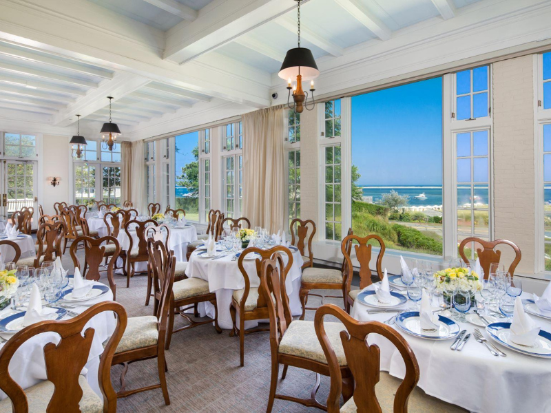 An elegant dining room set for an event, with round tables, white tablecloths, and wooden chairs, overlooking a scenic ocean view through large windows.