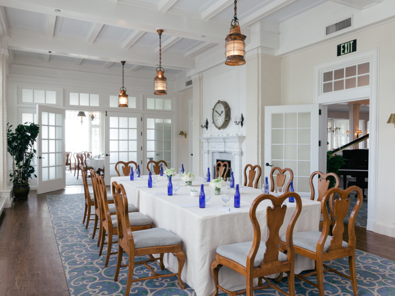A elegant dining room with a long table, wooden chairs, blue bottles, and a fireplace. The room is bright with large windows.