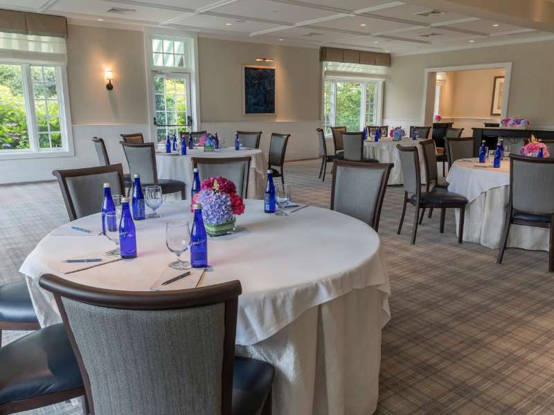 A well-lit room with round tables set for a gathering, featuring blue water bottles and floral centerpieces.