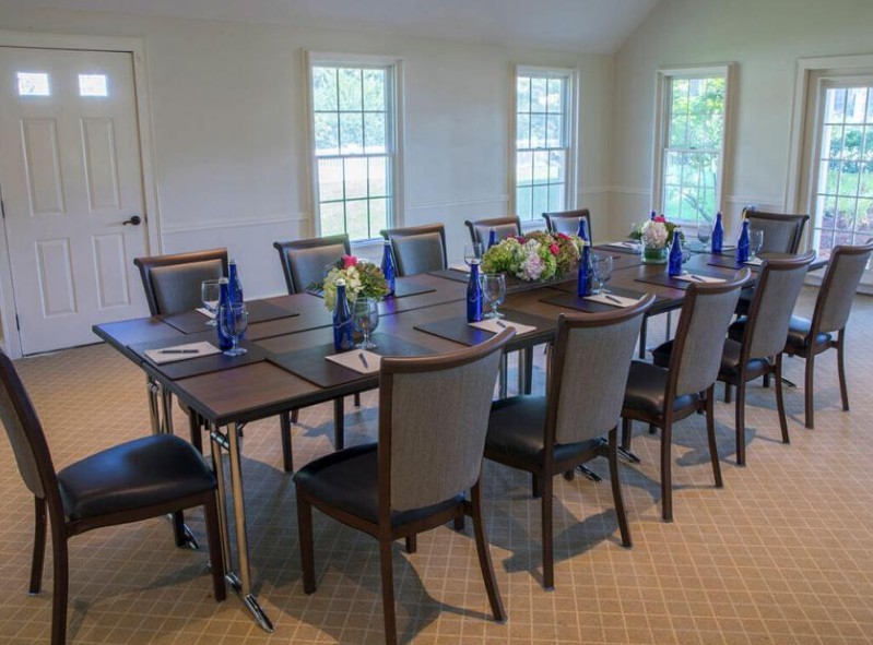 A well-lit meeting room with a large table, twelve chairs, blue bottles, notepads, and flower arrangements decoratively placed.