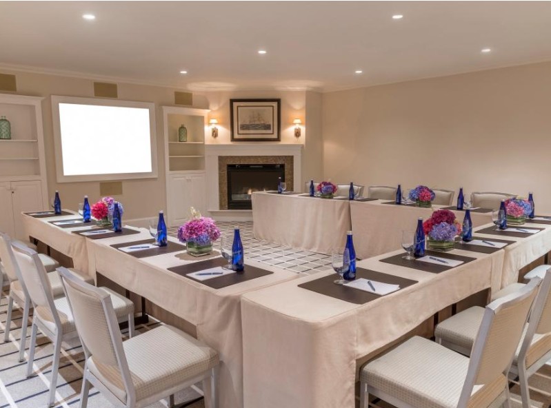 A conference room with a U-shaped table arrangement, notepads, pens, blue water bottles, floral centerpieces, and a presentation screen.