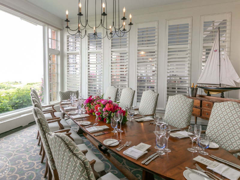 An elegant dining room with a long table, floral centerpiece, plates, glassware, and a model sailboat in a bright space.