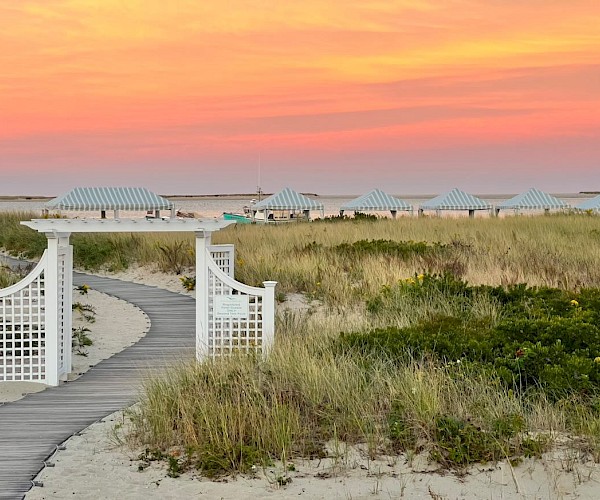 A picturesque beach scene with a wooden path, white gate, grassy dunes, striped tents, and a vibrant sunset sky.