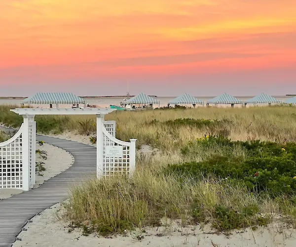 A picturesque beach scene with a wooden path, white gate, grassy dunes, striped tents, and a vibrant sunset sky.