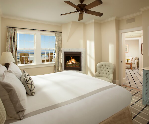 A cozy bedroom with a bed, fireplace, armchair, and window view, leading to a sitting area with a sofa.