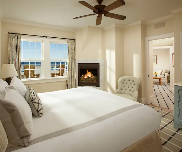 A cozy bedroom with a bed, fireplace, armchair, and window view, leading to a sitting area with a sofa.