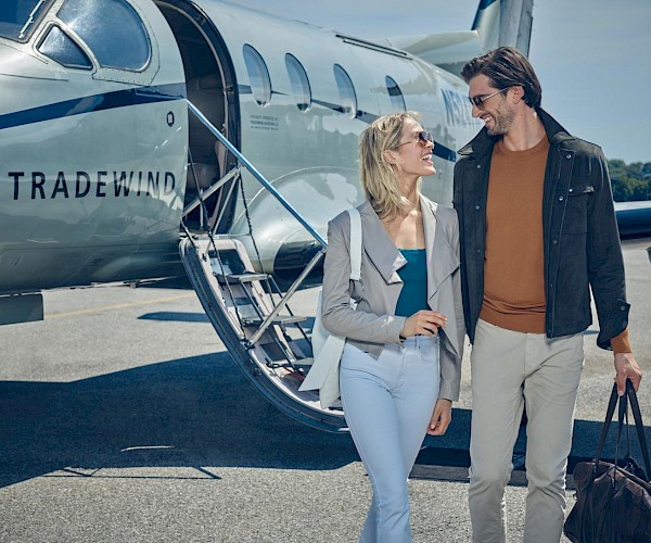 A couple walks away from a small plane labeled "Tradewind," smiling and carrying a bag on a sunny day.