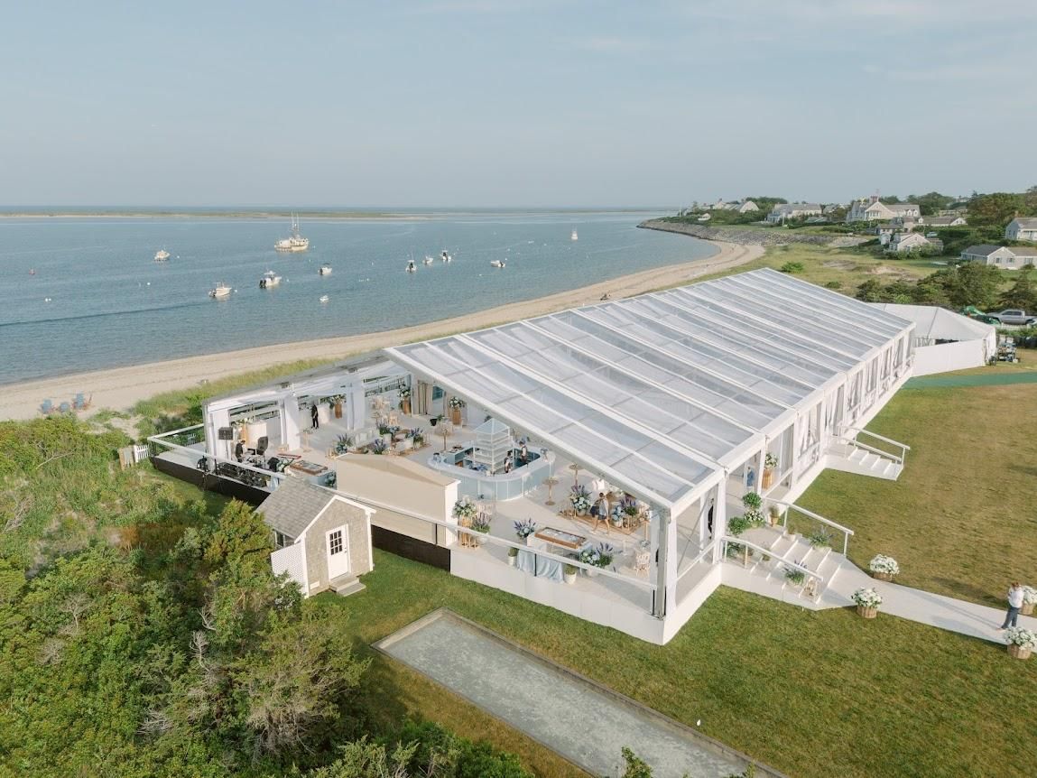 A large white tented venue by the water with boats in the distance, surrounded by greenery and adjacent to a sandy beach.