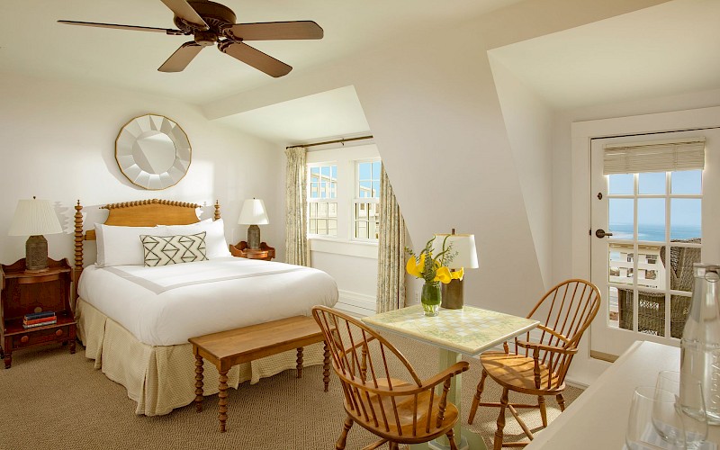 A cozy bedroom featuring a bed, two chairs, a table with flowers, and a view of the ocean through a glass door and window.