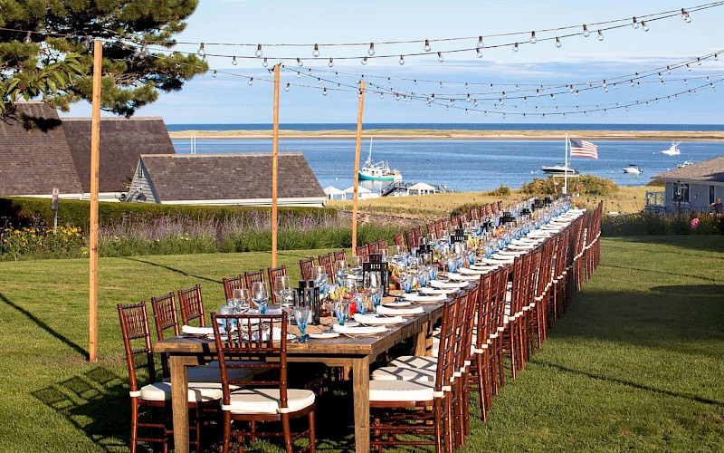 A long outdoor dining table with chairs is set on a lawn by the sea, decorated with lights and ready for an event or celebration.